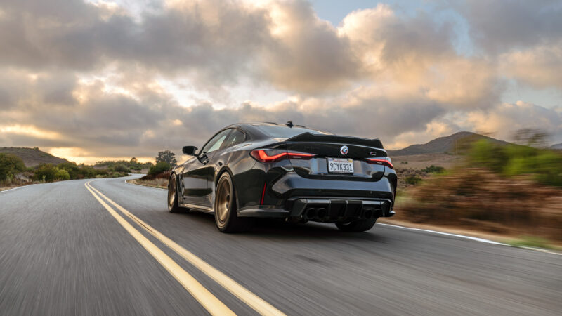 A black sportscar driving on a winding road, against a backdrop of cloudy sky and hills.