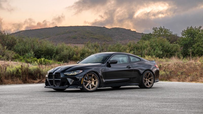 A black sports car parked on a paved surface with a backdrop of hills and a cloudy sky at sunset.