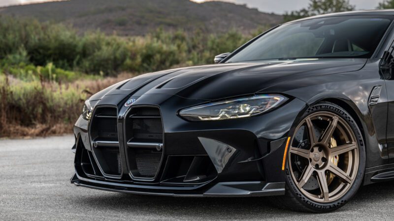 Close-up of a sleek black sports car with gold alloy wheels, parked on a road with greenery in the background.