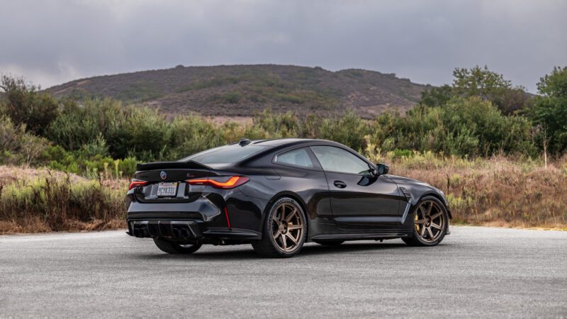 A black sports coupe with dark bronze wheels is parked on a paved area, with green hills and a cloudy sky in the background.