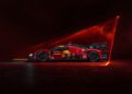 A red Ferrari 499P Hypercar adorned with various logos is illuminated in dramatic lighting, set against a dark backdrop with striking red lighting effects, showcasing the sleek lines of its 2025 livery.