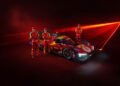 The 499P Hypercar with its striking 2025 livery in red and yellow is on display, flanked by three individuals in matching Ferrari racing suits and caps, set against a vibrant red laser backdrop.