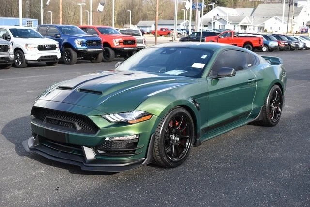 A green sports car with a black racing stripe, a unique creation among custom cars, is parked on a dealership lot, surrounded by other vehicles.