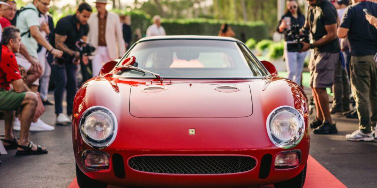 A red vintage Ferrari 250 LM sports car, often hailed as the Best of the Best, is parked on a red carpet, surrounded by a crowd of people snapping photos at the prestigious Cavallino Classic event.