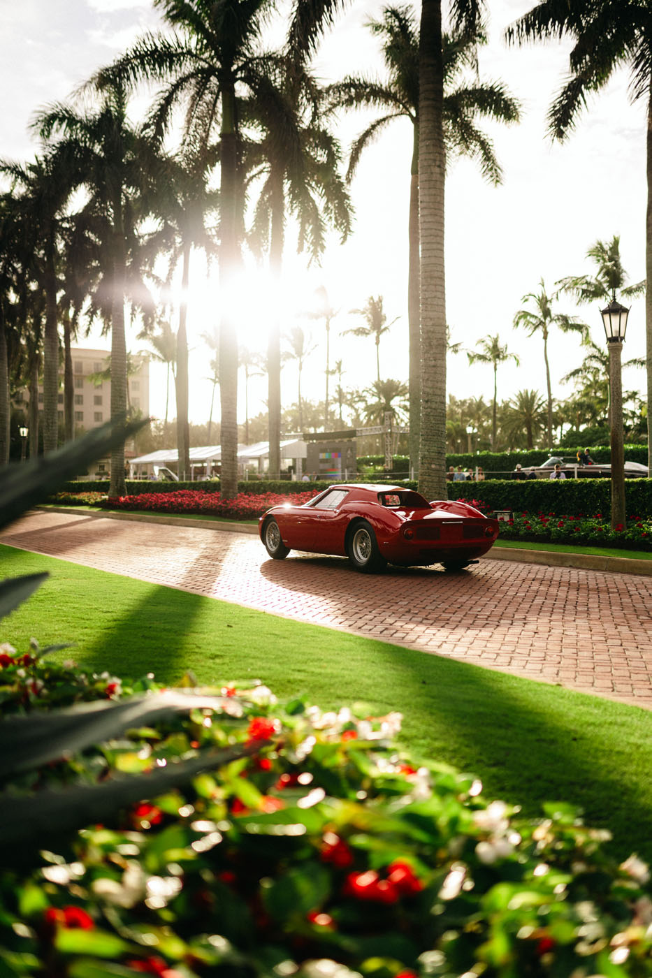 A red convertible Ferrari 250 LM cruises along a brick road lined with palm trees and flowers at the Cavallino Classic. Sunlight filters through the trees, casting a golden glow on this Best of the Best journey.