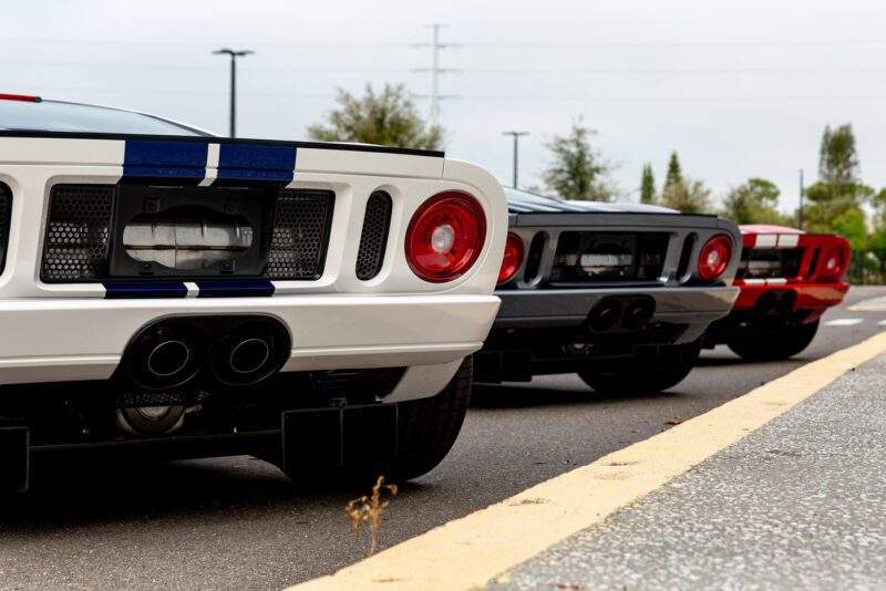 Tampilan belakang tiga mobil sport yang diparkir dalam satu baris, menampilkan lampu belakang bundar yang khas dan pipa knalpot ganda. Ford GT 2006 yang ramping menonjol di antara mereka, desain ikoniknya membuat para penonton ingin memenangkan tawaran.