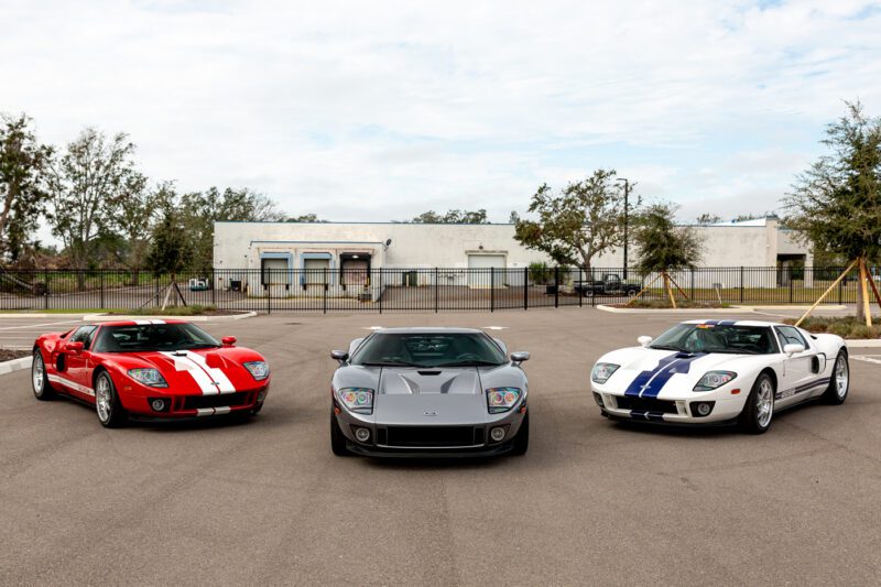 Tiga mobil Ford GT yang menakjubkan dari tahun 2006 diparkir berdampingan di area beraspal; Dari kiri ke kanan: merah dengan garis -garis putih, perak dengan garis -garis hitam, dan putih dengan garis -garis biru. Pohon dan bangunan membingkai adegan sempurna yang akan ditawar oleh kolektor mana pun.