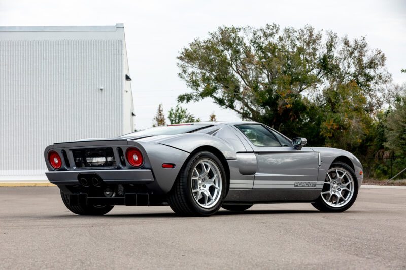 Mobil sport Ford GT 2006 yang ramping dengan tubuh yang ramping dan lampu ekor melingkar terletak dengan anggun di area beraspal. Di latar belakang, pohon dan bangunan besar melengkapi adegan yang menarik ini, mengundang Anda untuk mempertimbangkan memenangkan tawaran pada Ford GT yang sangat indah.