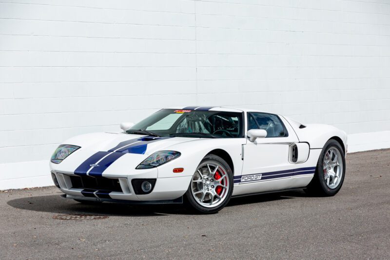 Ford GT 2006, mobil sport putih dengan garis -garis balap biru yang mencolok, diparkir di permukaan beraspal di depan dinding putih yang murni. Jangan lewatkan kesempatan Anda untuk memenangkan penawaran untuk kendaraan legendaris ini.