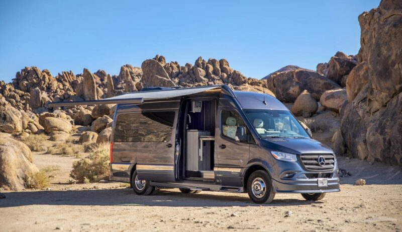 A gray camper van, a unique creation with an extended awning, is parked on a rocky desert landscape under a clear blue sky.