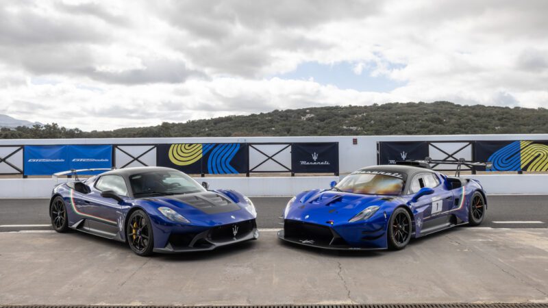 Two blue sports cars are parked on a track. One has racing decals and the number 41, the other has the number 17. Hills and cloudy skies form the background.