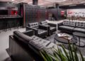 Sleek lounge with black leather sofas, round tables, and a wall-mounted tire display reminiscent of the Circuit de Barcelona-Catalunya. A bar area with stools is in the background, set against a modern industrial design.