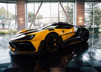 Yellow and black sports car displayed indoors on a reflective black tiled floor, with large windows and trees visible outside.