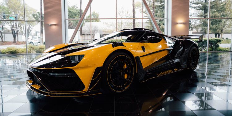Yellow and black sports car displayed indoors on a reflective black tiled floor, with large windows and trees visible outside.