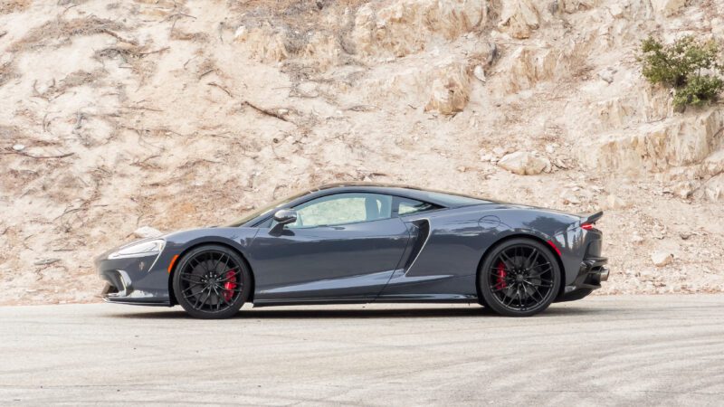 A sleek, gray sports car with black rims and red brake calipers is parked on a road, with a rocky hill in the background.