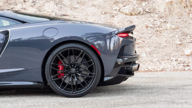 The rear section of a gray sports car with a visible taillight, exhaust, and black alloy wheel, parked on a road with a rocky background.