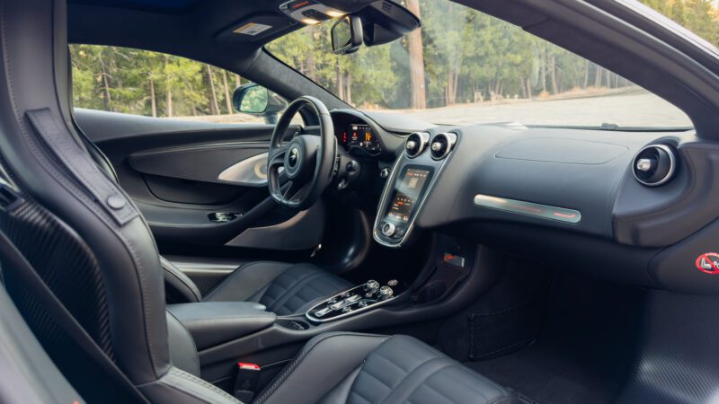 Interior of a modern sports car with black leather seats, digital dashboard, and central touchscreen. Forest visible through windows.