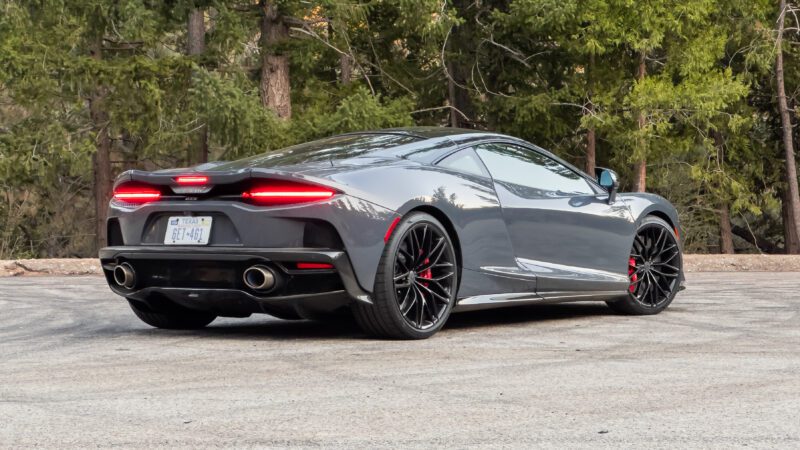 A sleek, gray sports car with tinted windows is parked on a road surrounded by trees. The car features red brake calipers and dual exhausts.