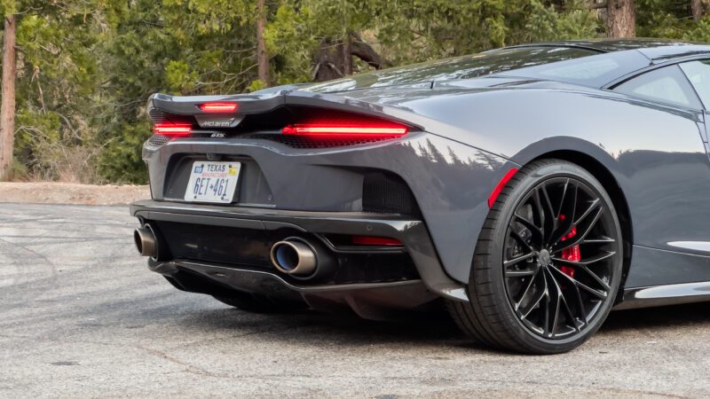 Rear view of a gray sports car with Texas license plate, showing dual exhaust pipes and black alloy wheels, parked on a paved surface with trees in the background.