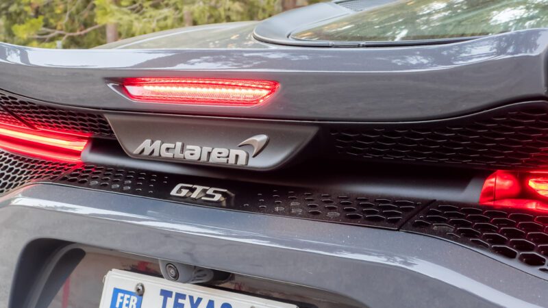 Close-up of the rear end of a grey McLaren GT with visible taillights, exhaust vents, and a Texas license plate.