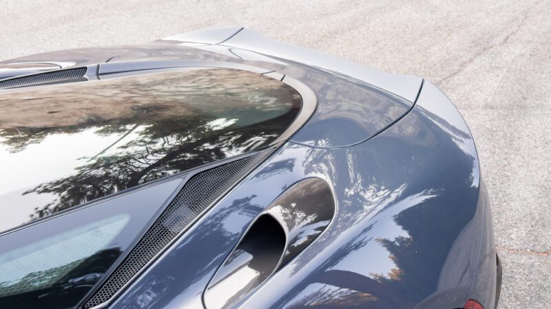 Close-up of a sleek car's rear with a glossy finish reflecting trees. The design includes aerodynamic elements and vents.