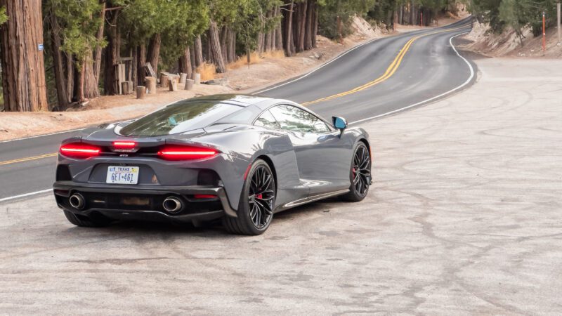 A gray sports car with a Texas license plate is parked on a roadside, surrounded by tall trees.