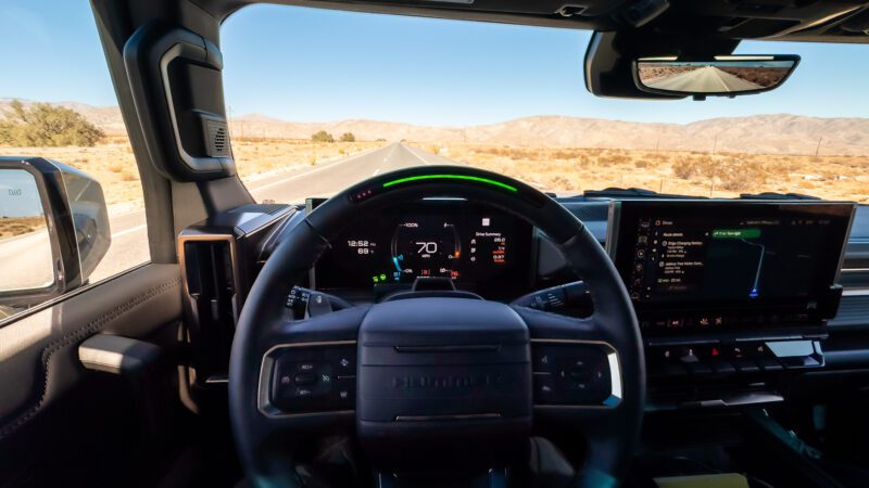 Interior view of a vehicle with autonomous driving features, showing a steering wheel, dashboard with digital display, and road through the windshield in a desert landscape.