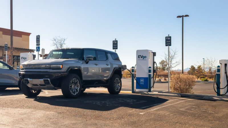 A silver electric SUV is parked at an EVgo charging station in a parking lot under clear skies.