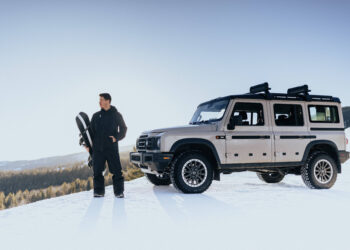 A person stands next to a beige SUV on a snowy hill, inspired by The Snow League, holding a snowboard under a clear blue sky.