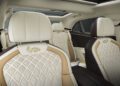 Experience the exquisite elegance of a Bentley's interior, part of the Bespoke Collections by Mulliner, featuring quilted leather seats in beige and white with intricate stitching and a dragon emblem on the headrests. A sunroof graces this luxurious ride.