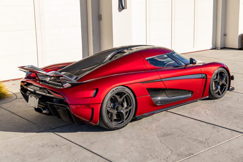 A red Koenigsegg Regeras sports car is parked on a concrete driveway in front of a white garage door, exuding extreme elegance and power.