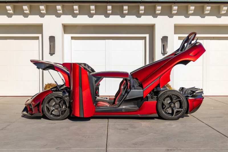 A red Koenigsegg Regera sports car with its doors, hood, and rear engine cover open sits on a driveway in front of a garage, displaying its extreme design and engineering marvel.