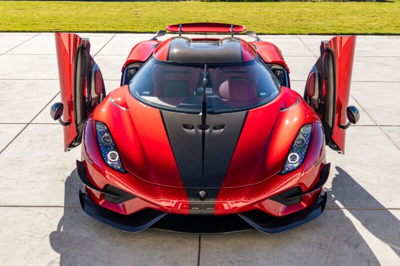 A red sports car, reminiscent of the extreme Koenigsegg Regera, with vertically open doors, is parked on a concrete surface. It boasts a sleek design and a black central stripe, while grass peeks from the background.