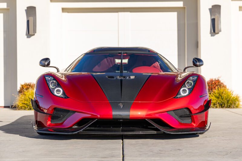 The sleek red Koenigsegg Regera with black accents, viewed head-on, sits parked in front of a modern white building, showcasing its extreme design.