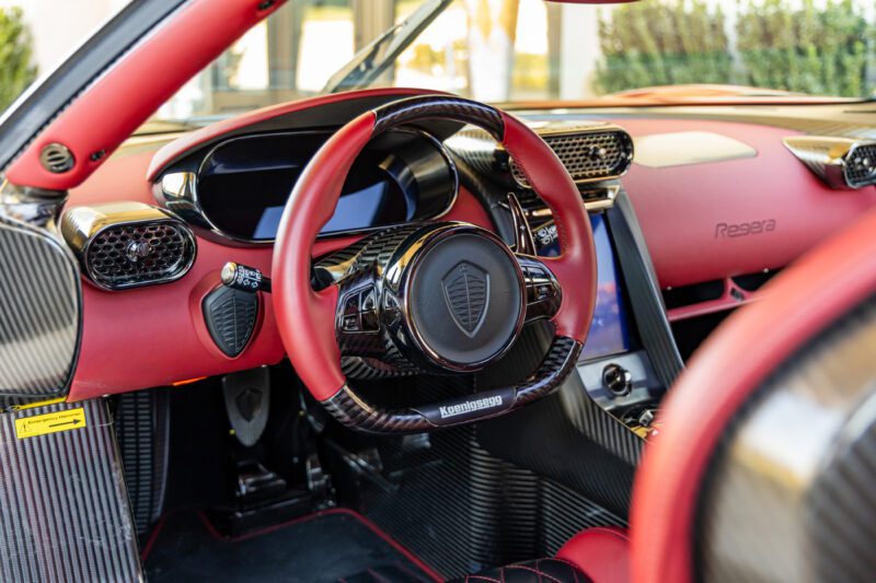 The interior of this extreme luxury sports car boasts a Koenigsegg Regera steering wheel, vibrant red leather accents, a sleek digital display, and sophisticated carbon fiber details.