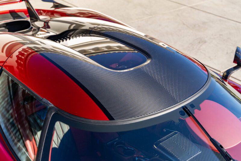 A close-up of a Koenigsegg Regera car roof showcases a sleek carbon fiber pattern with extreme red and black accents, reflecting clear skies above.