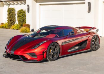 A Koenigsegg Regera is parked outside a modern building with white garage doors and lush plants nearby, showcasing its extreme car design in striking red and black.
