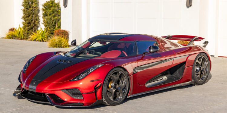 A Koenigsegg Regera is parked outside a modern building with white garage doors and lush plants nearby, showcasing its extreme car design in striking red and black.