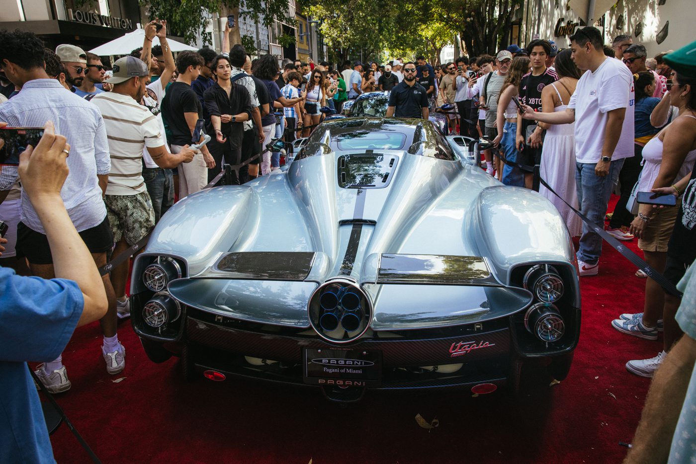 At the Miami Concours 2025, a crowd gathers around a sleek, silver sports car displayed on a red carpet. Attendees of this Supercar Art Event take photos and admire the vehicle's design under leafy trees.