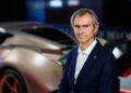 A man in a dark suit stands beside a Hispano Suiza, its sleek design features and vivid lighting reflecting an aura of sophistication, evoking images of the Circuit de Barcelona-Catalunya.
