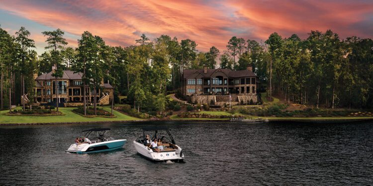 Two boats glide across a lake near upscale lakeside houses in the renowned Reynolds Lake Oconee community, surrounded by lush trees at sunset. The vibrant, orange-pink sky above adds to the charm of what many consider the South's Best Lake.