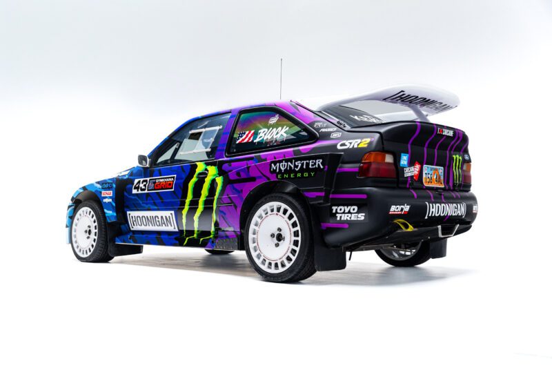 Blue and purple rally car with Monster Energy and Hoonigan logos, white wheels, and a rear spoiler, parked against a white background.