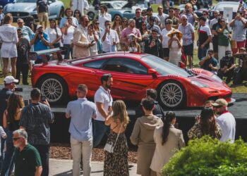 A red sports car is displayed on a platform at duPont REGISTRY's Ultimate Weekend Guide event, with a crowd of people taking photos and admiring the sleek vehicle.