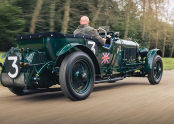 A vintage green Bentley Speed Six from the Continuation Series, with the number 3 and a British flag on the side, is being driven on a road by a man. The classic car features spoked wheels and a rear-mounted spare tire.