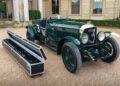 A vintage green Bentley Speed Six Continuation Series race car is parked in front of a historic building, with a long open case containing detached parts beside it, each component echoing a special story from the past.