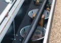 Close-up of a long, black-barrelled item with metal grilles, reminiscent of the Bentley Speed Six, secured inside an open, metallic-framed container on a gravel surface.