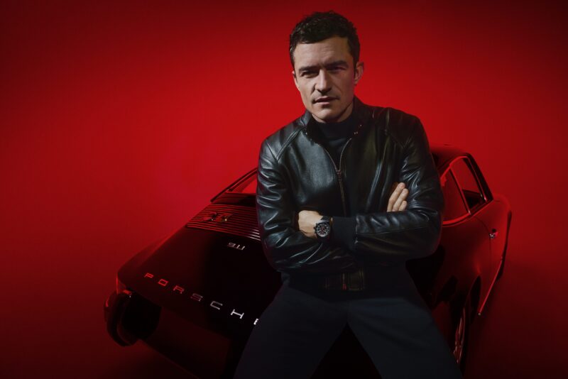 Orlando Bloom, mengenakan jaket kulit, berdiri dengan lengan yang disilangkan di depan Porsche hitam di bawah pencahayaan merah, mewujudkan perannya sebagai duta merek untuk desain Porsche.