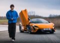 A person in a blue jacket casually walks near an orange McLaren sports car with its door open, parked on a paved area.