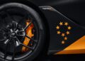 Close-up of a black McLaren 720S wheel with orange brake caliper and logo. The side panel showcases a circular pattern of orange stars, partially cut off by the wheel arch, echoing the sleek design elements seen in their MCL38 F1 Car.