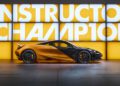A sleek orange and black sports car, reminiscent of the McLaren 750S and Artura editions, is parked indoors against a brightly lit wall displaying the words "CONSTRUCTOR CHAMPION.